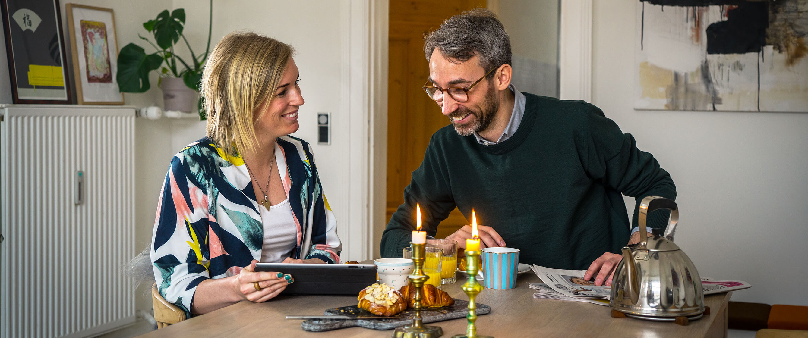 Billede, der viser to, der sidder og spiser morgenmad. De kigger på en tablet, der viser dem abonnementsløsningen: MobilePay Subscriptions. 